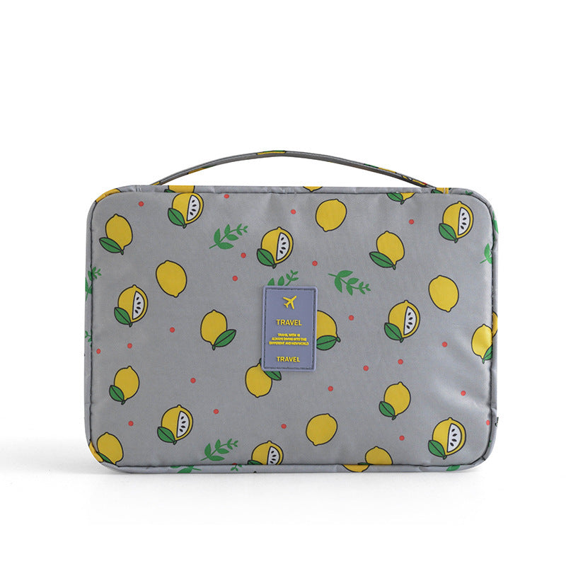 Gray travel cosmetic bag with a yellow lemon print, top handle, and central "TRAVEL" logo patch.