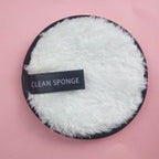 Round white plush makeup remover puff with a black trim and "CLEAN SPONGE" label on a pink background.