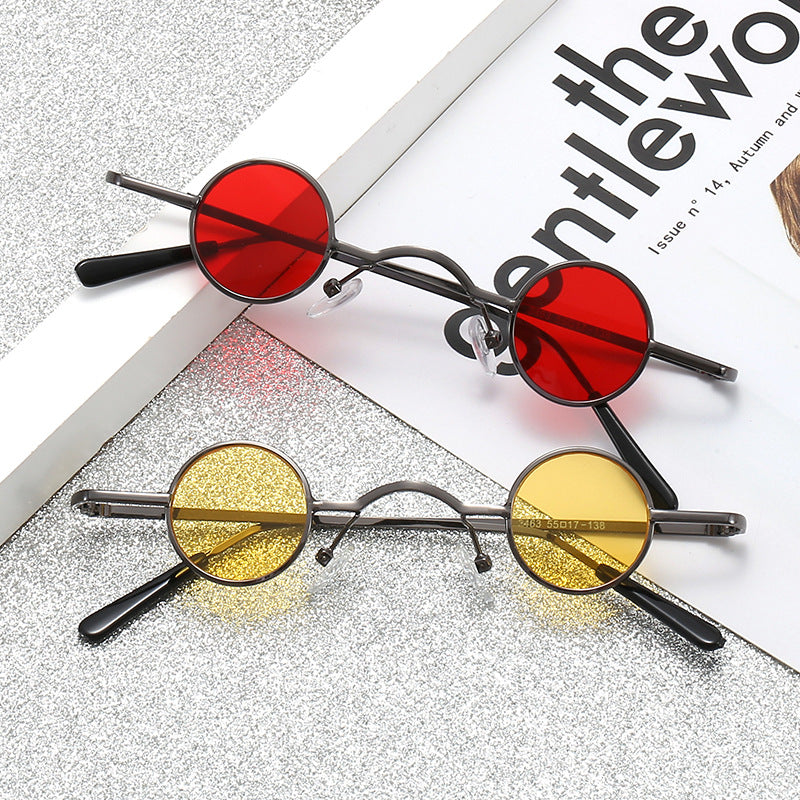 Two pairs of retro sunglasses with thin, dark metal frames, round red and yellow lenses on a glittery surface.