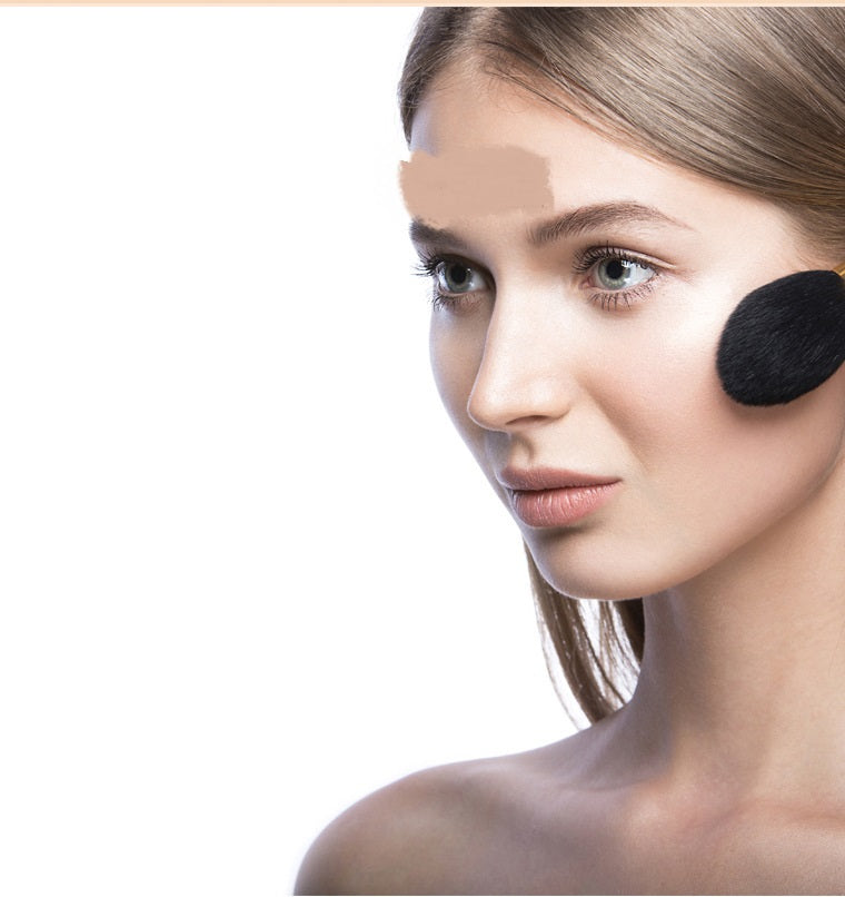 A woman having her cheek brushed with a black-bristled makeup brush against a plain white background.