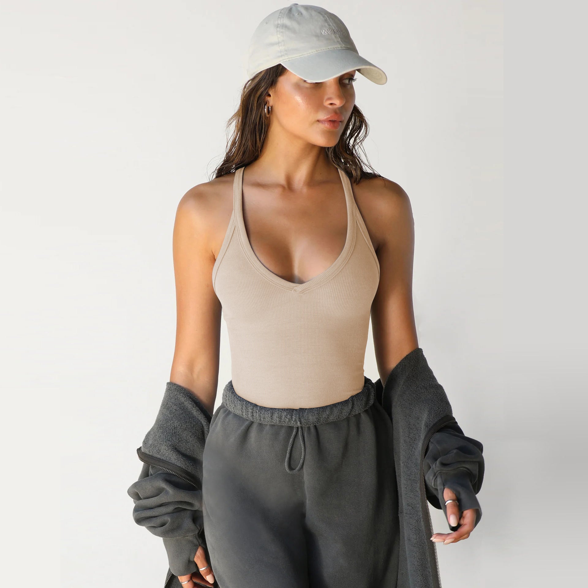 A woman wears a beige, ribbed, V-neck racerback tank top with a grey baseball cap and charcoal-colored sweatpants.