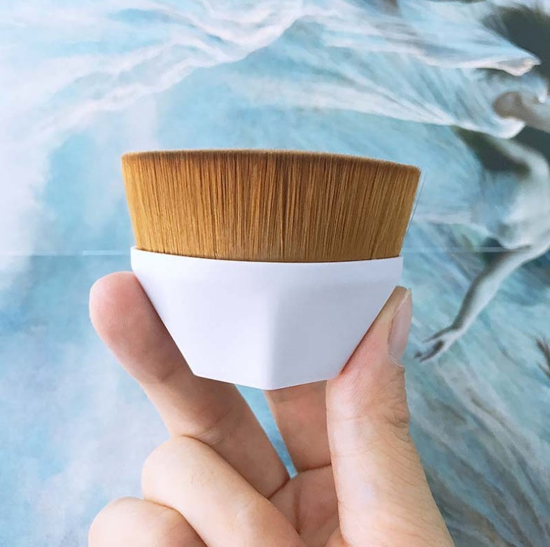 Hand holding a flat-top makeup brush with synthetic brown bristles and a white plastic ergonomic handle.