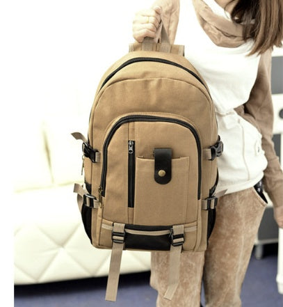 A person holds a khaki canvas backpack with black leather accents, multiple zippered pockets, and adjustable side straps.
