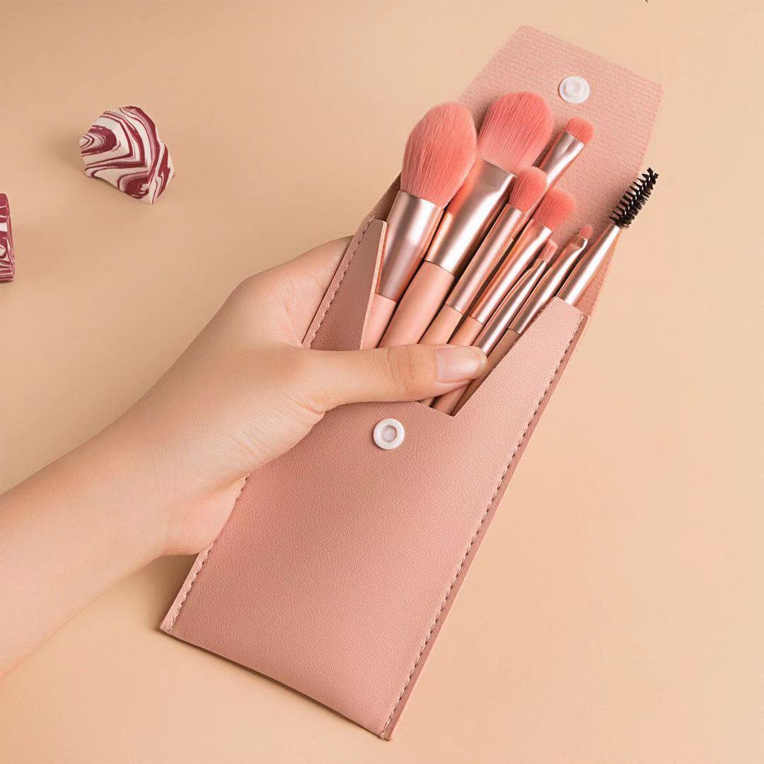 Hand holding a set of 8 rose gold portable makeup brushes in a matching pink faux-leather snap-button travel case.