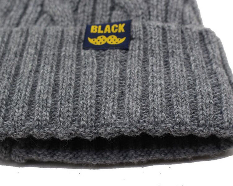 Close-up of a grey ribbed knit winter beanie with a small navy tag featuring yellow "BLACK" text and a mustache icon.