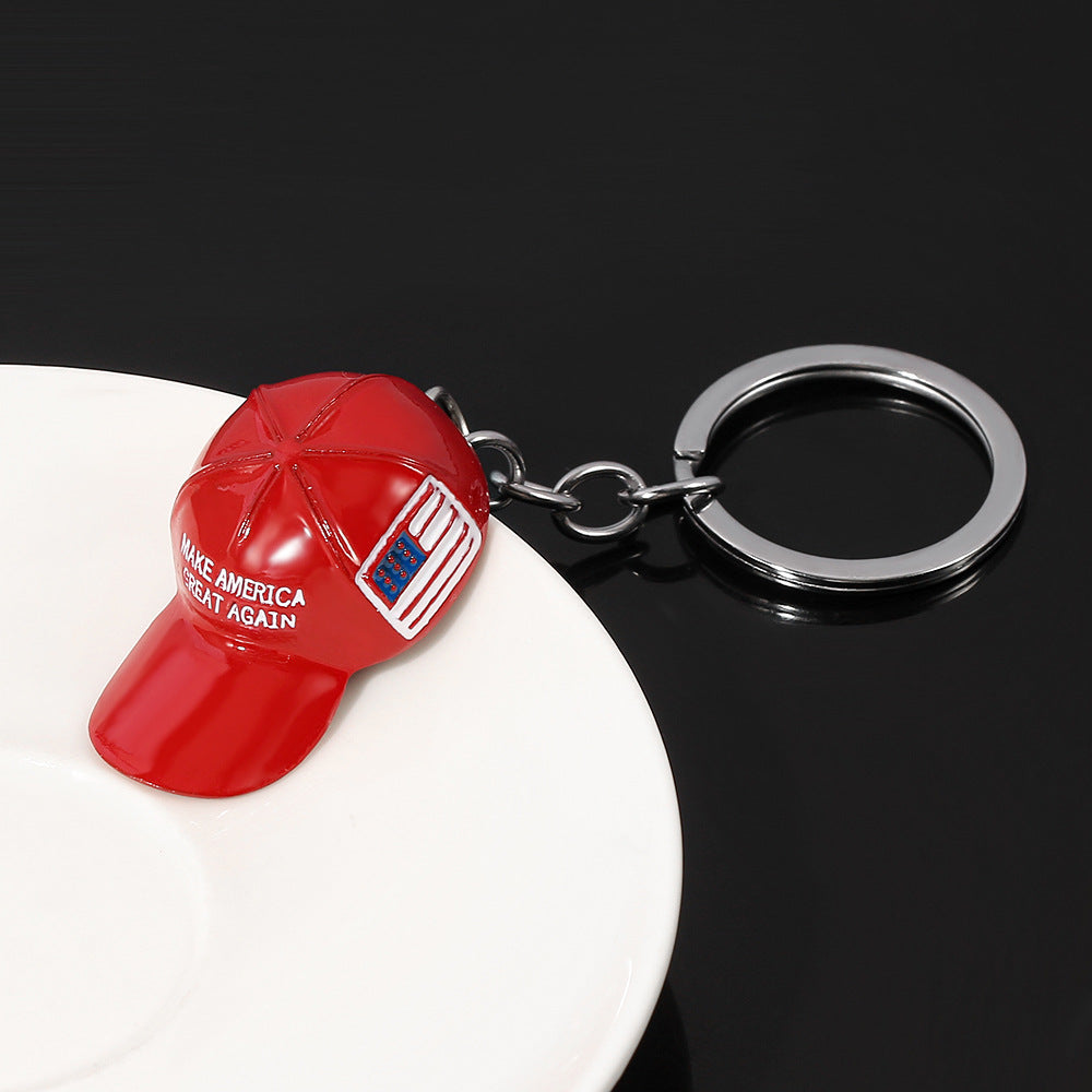Red enamel hat keychain with "MAKE AMERICA GREAT AGAIN" text, a flag emblem, and a metal key ring on a white plate.