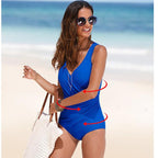 Woman on beach wearing a bright blue, V-neck, ruched one-piece swimsuit, carrying a textured white tote bag.