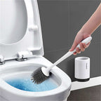 A person cleans a white toilet bowl with a gray silicone brush featuring a long white handle and matching holder.