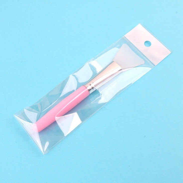 A pink-handled silicone face mask brush with a metallic gold ferrule, packaged in a clear plastic bag on a blue background.