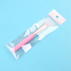 A pink-handled silicone face mask brush with a metallic gold ferrule, packaged in a clear plastic bag on a blue background.