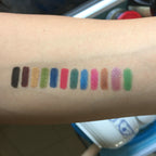 Arm showing 12 swatches of colorful, glittery, and metallic eyeliner pencil makeup in various bright shades.