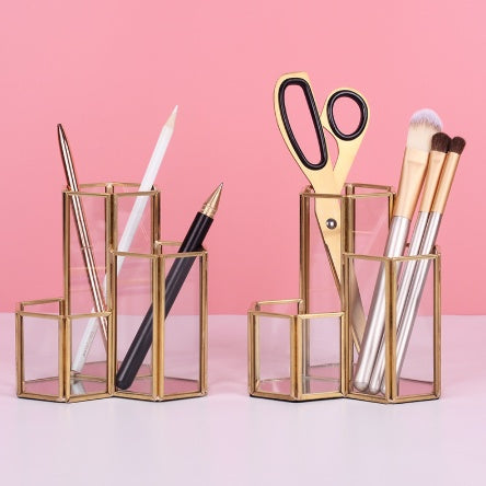 Two hexagonal clear glass and gold-trimmed makeup brush holders holding stationery and brushes on a pink background.