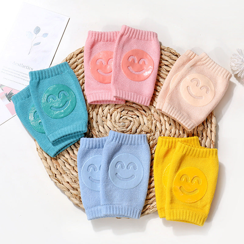 Multi-colored terry cloth baby knee pads with silicone smiley face grips, arranged on a round woven mat.