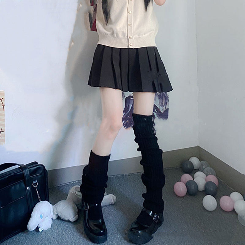 Person wearing black ribbed, pile-style knitted leg warmers over black shoes with a pleated skirt.