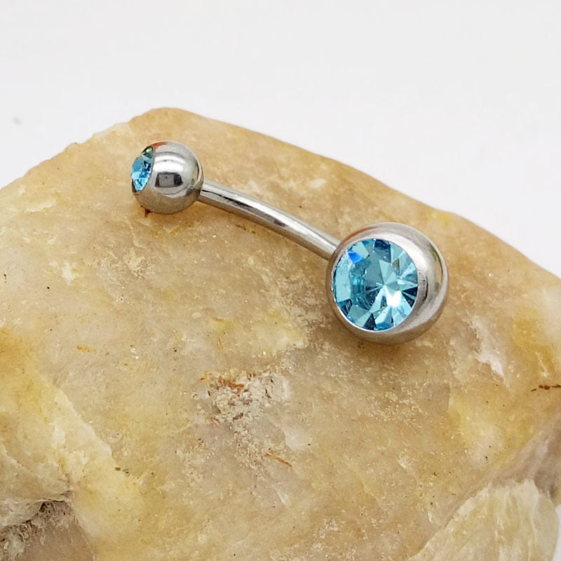 Titanium steel belly button ring featuring two blue crystal gems, resting on a textured, light-colored stone.