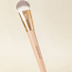 Soft flat-head makeup brush with dense grey/white bristles, gold ferrule, and a beige handle. Neutral cream background.