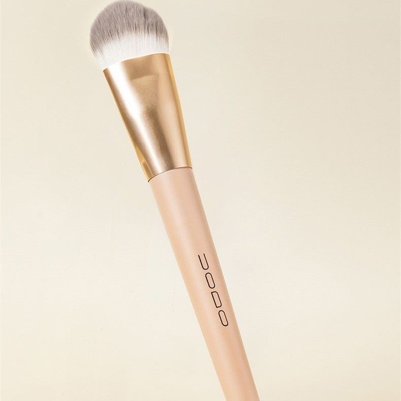 Soft flat-head makeup brush with dense grey/white bristles, gold ferrule, and a beige handle. Neutral cream background.