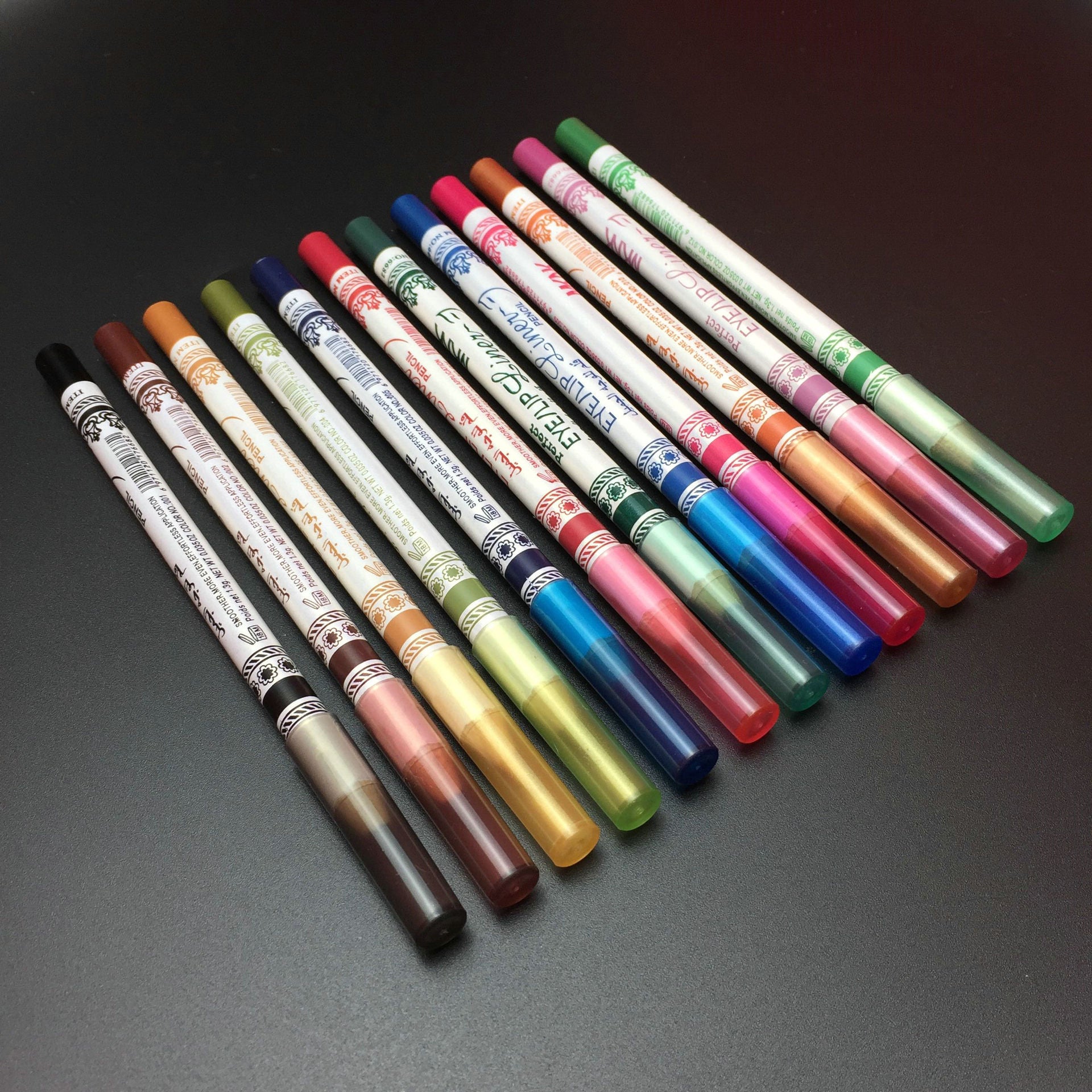 A set of 12 vibrant, glitter-finish eye and lip liner pencils arranged diagonally on a dark surface.