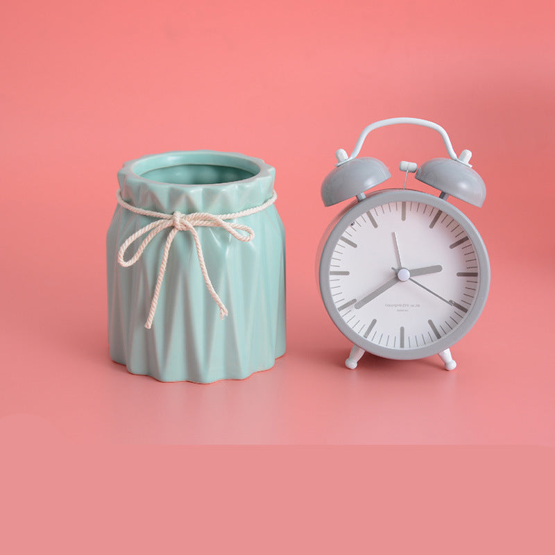 Mint green fluted ceramic makeup brush holder with a white rope bow tie next to a grey alarm clock on a pink surface.