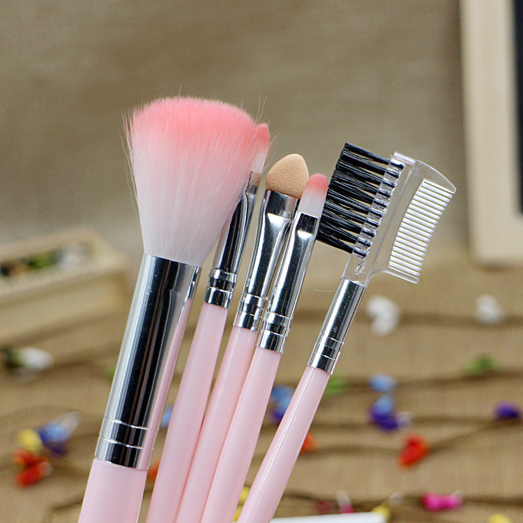 Five makeup brushes with light pink handles, silver ferrules, and a mix of soft bristles, sponge, and lash comb/brush.