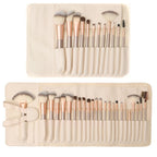 Set of 24 makeup brushes with champagne gold handles and synthetic white hair, stored in a cream-colored roll-up case.