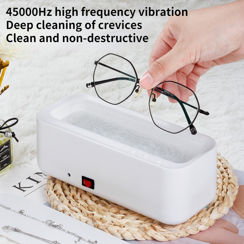 A white, rectangular ultrasonic glasses cleaner with a red power switch on a woven mat, with text about 45000Hz cleaning.