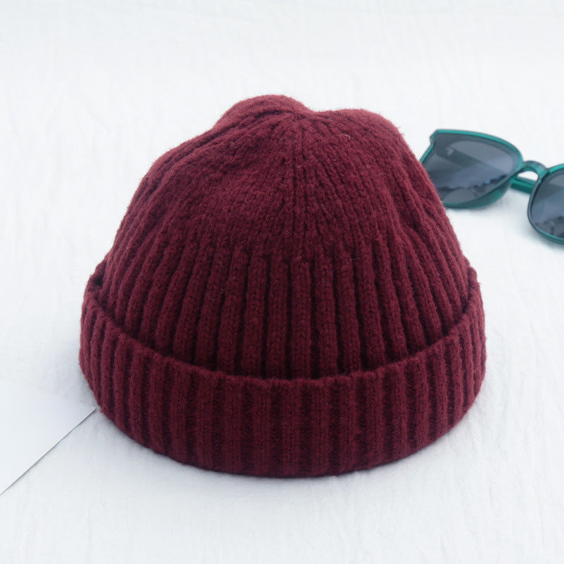 Maroon ribbed knit beanie with a folded cuff on a white background, next to dark-rimmed sunglasses.