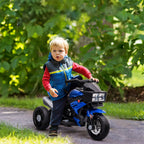 A young boy rides a blue plastic 3-wheeled electric motorcycle toy with headlights on a paved path outdoors.