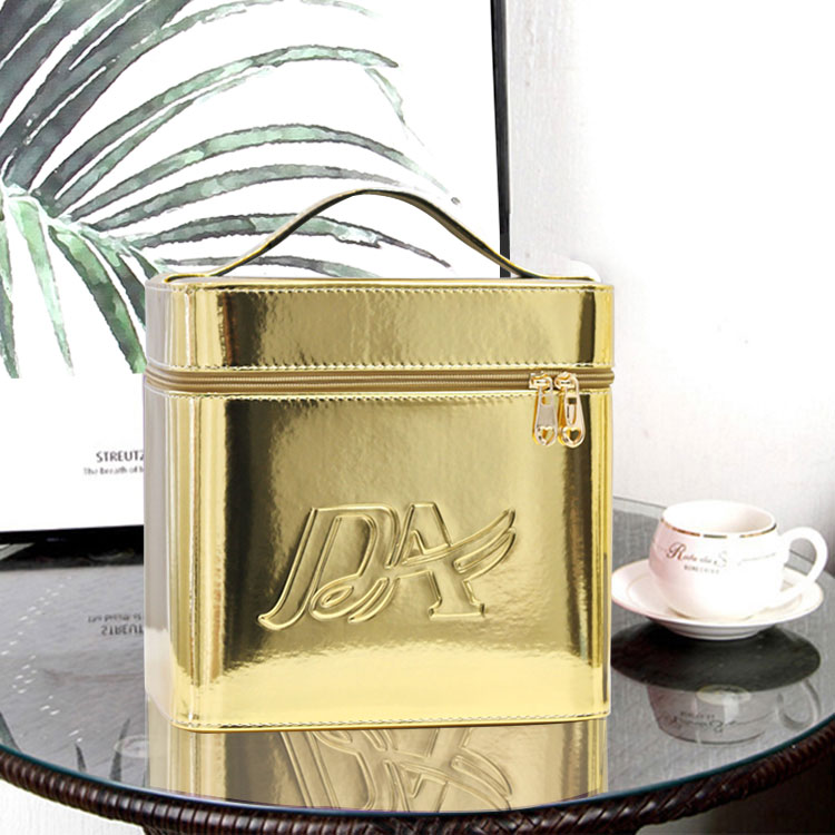 Metallic gold, rectangular portable makeup bag with top handle, zipper closure, and embossed logo on a glass table.