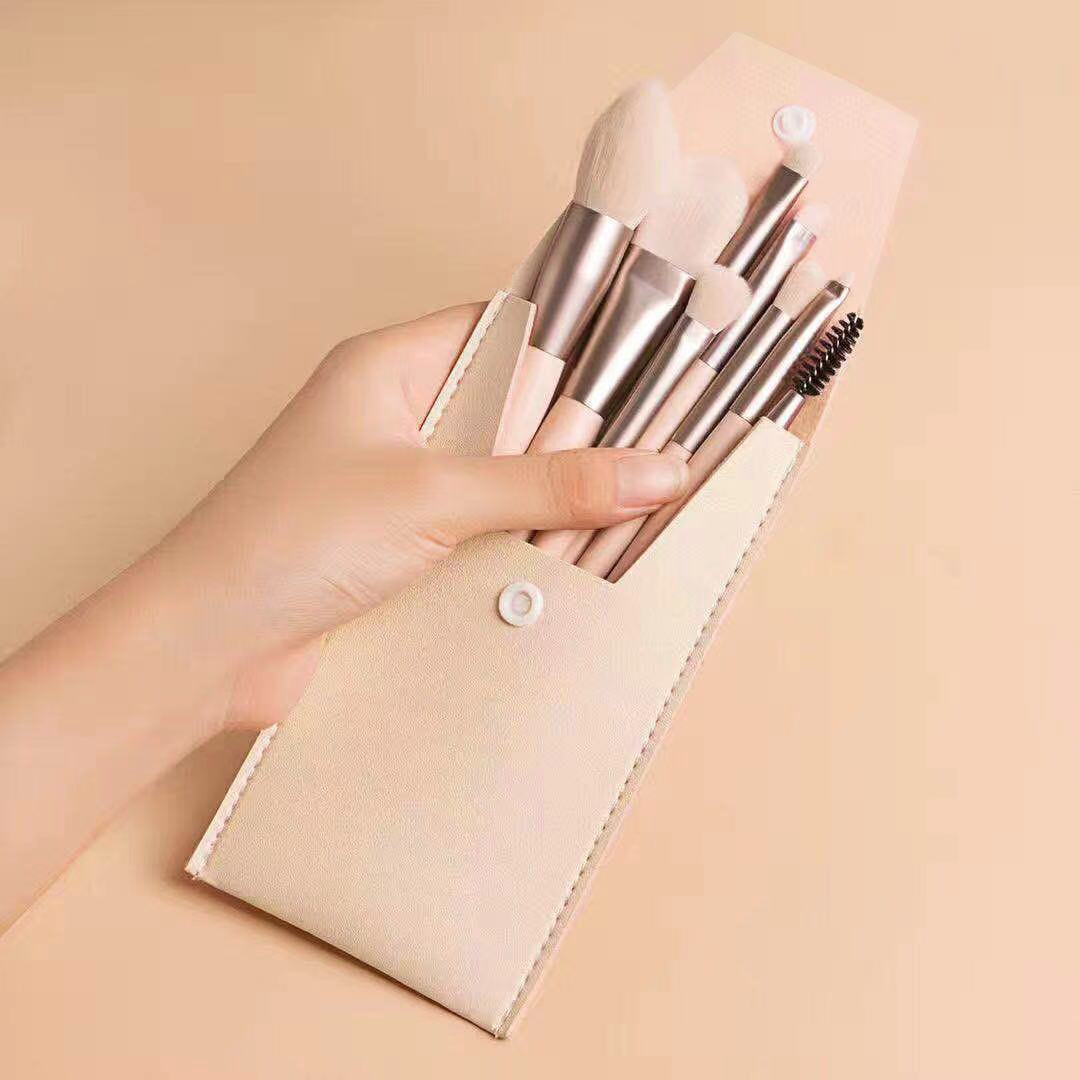 A hand holds a portable beige makeup brush set with metallic handles in a matching snap-button faux leather case.