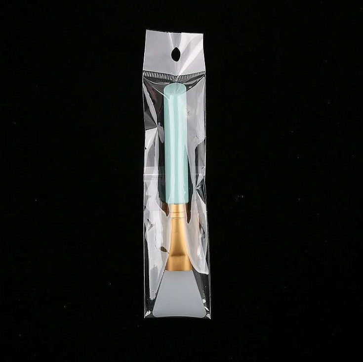 A silicone face mask brush with a light blue handle and gold ferrule, sealed in a clear plastic retail bag.