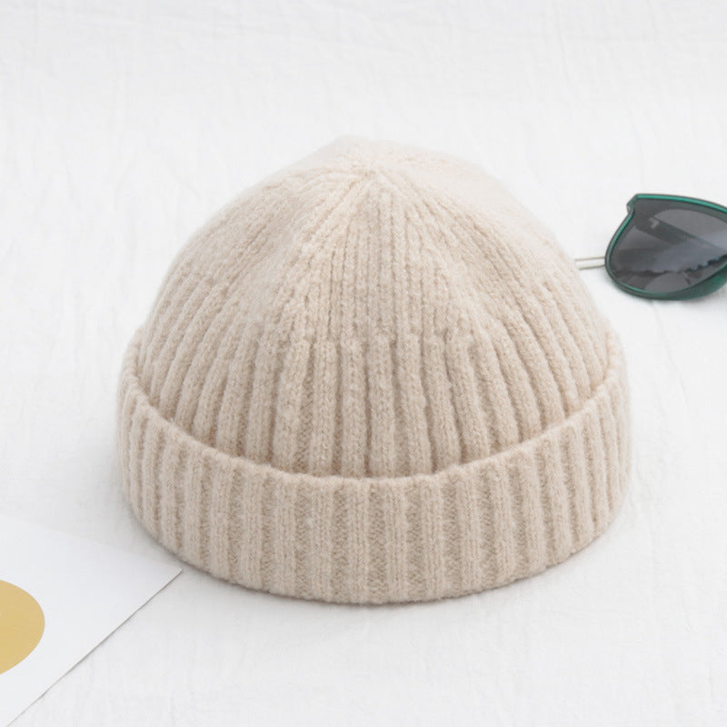 Beige ribbed knit beanie with a cuffed brim, placed on a white surface next to dark-framed sunglasses.