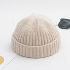 Beige ribbed knit beanie with a cuffed brim, placed on a white surface next to dark-framed sunglasses.