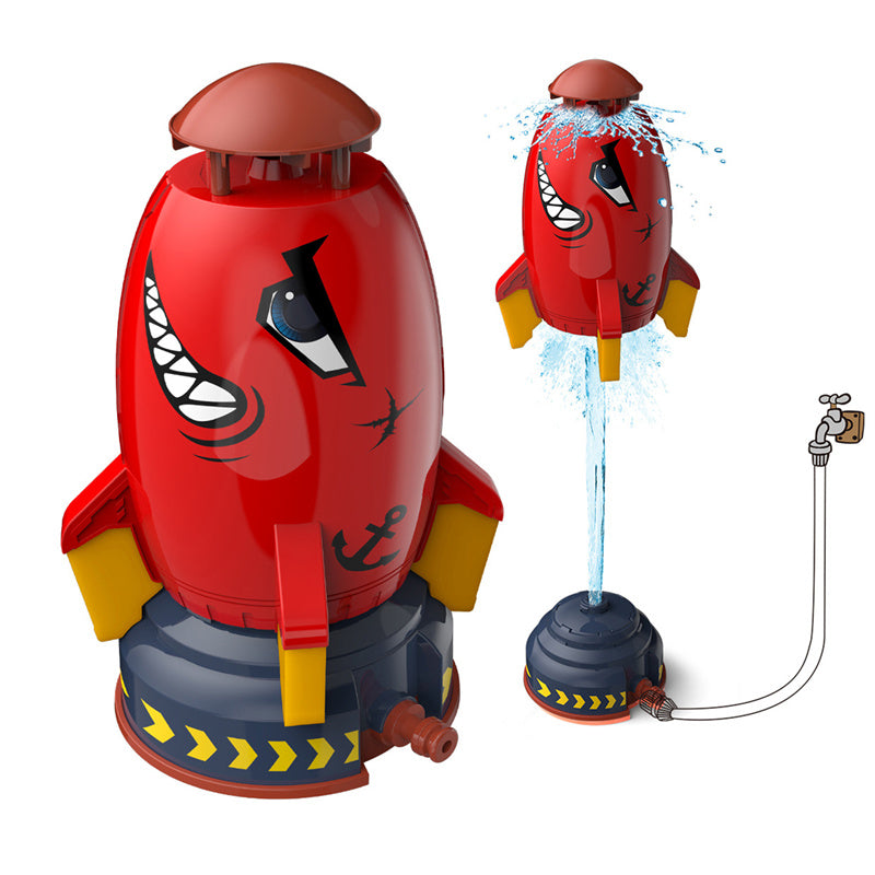 Red rocket sprinkler toy for kids with shark face, yellow fins, and blue base, connected to a garden hose for water lift.