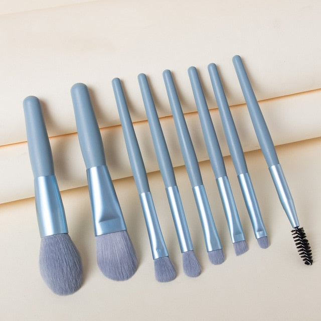 Eight portable makeup brushes with matte blue handles and matching blue bristles, arranged on a beige background.