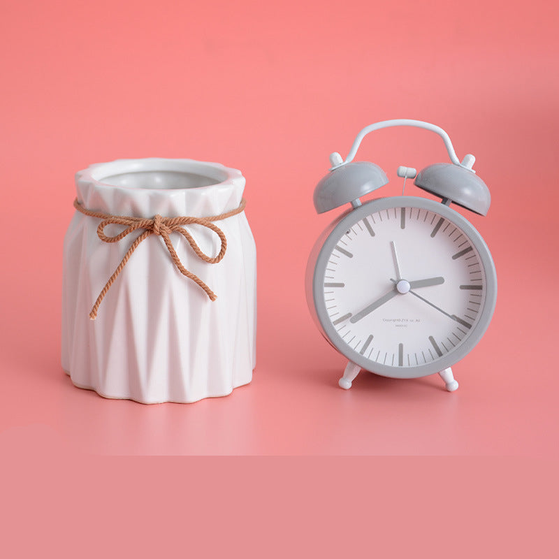 White ceramic pleated brush barrel with jute bow next to a grey alarm clock on a solid pink background.
