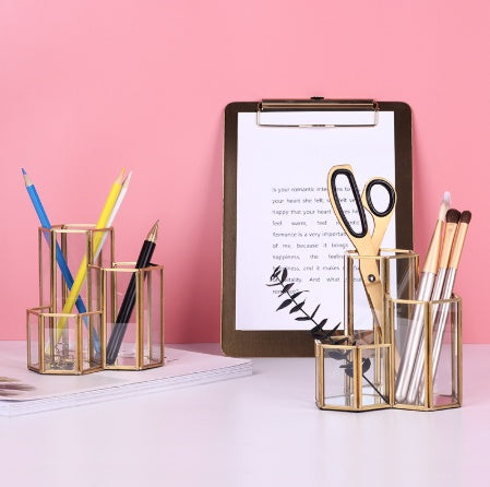 Two hexagonal gold-rimmed glass brush holders filled with makeup brushes, pens, and scissors on a white desk.