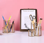 Two hexagonal gold-rimmed glass brush holders filled with makeup brushes, pens, and scissors on a white desk.