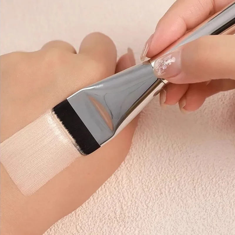 Ultra-thin foundation brush with silver handle applying a streak-free layer of liquid makeup on a hand.