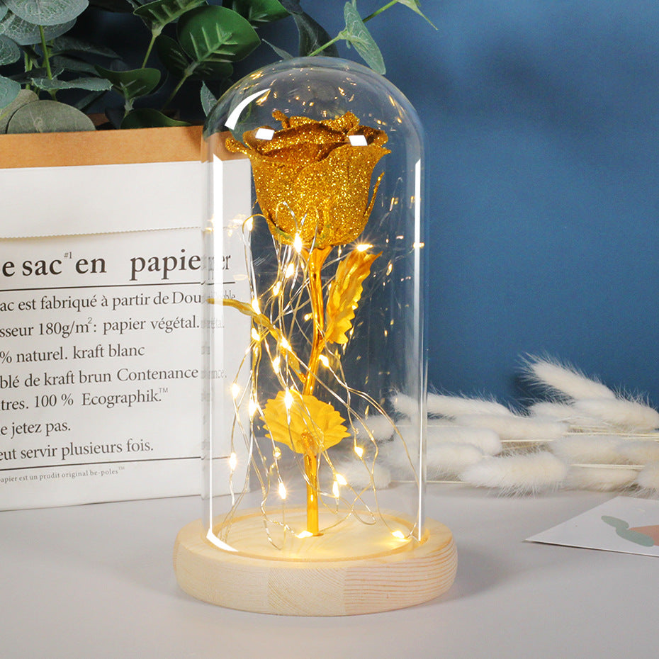A glittering gold rose inside a glass dome on a wooden base, illuminated by warm LED string lights.