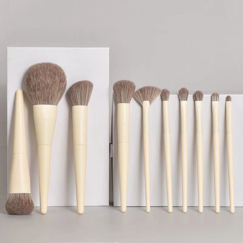 A set of nine makeup brushes with pale yellow handles and soft, light brown synthetic bristles against white blocks.