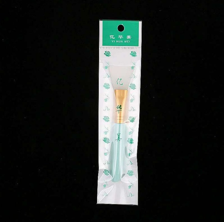 A silicone face mask brush with a gold ferrule and light blue handle, packaged in a branded plastic sleeve.