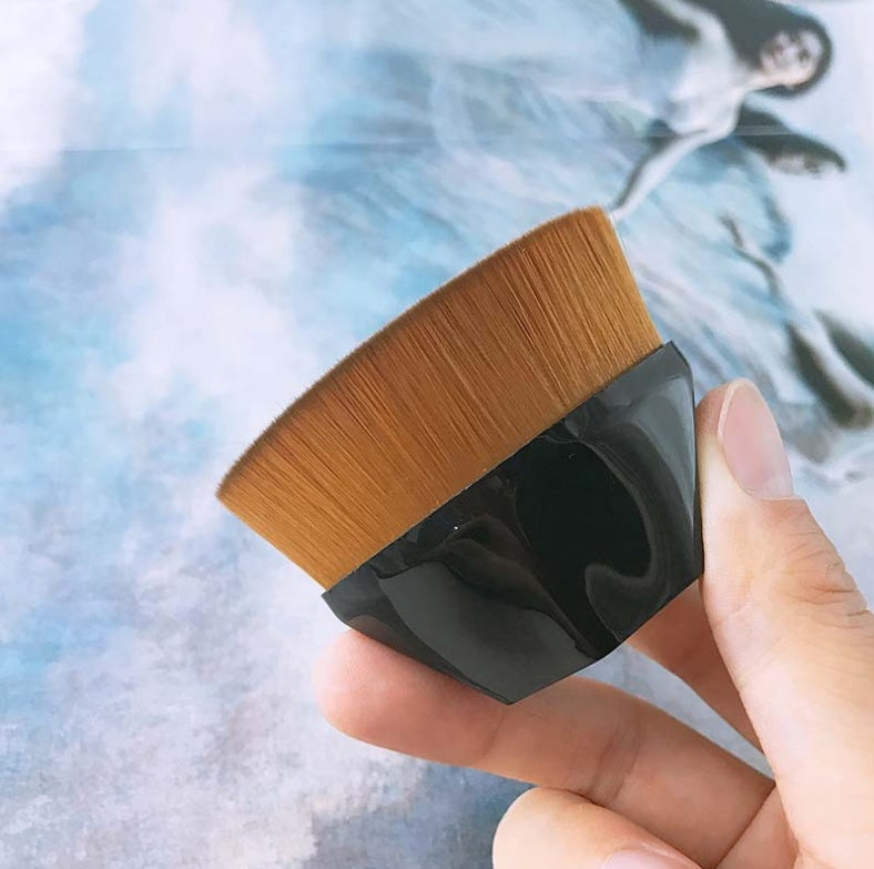 A hand holds a flat-top makeup brush with dense brown synthetic bristles and a glossy, faceted black plastic handle.