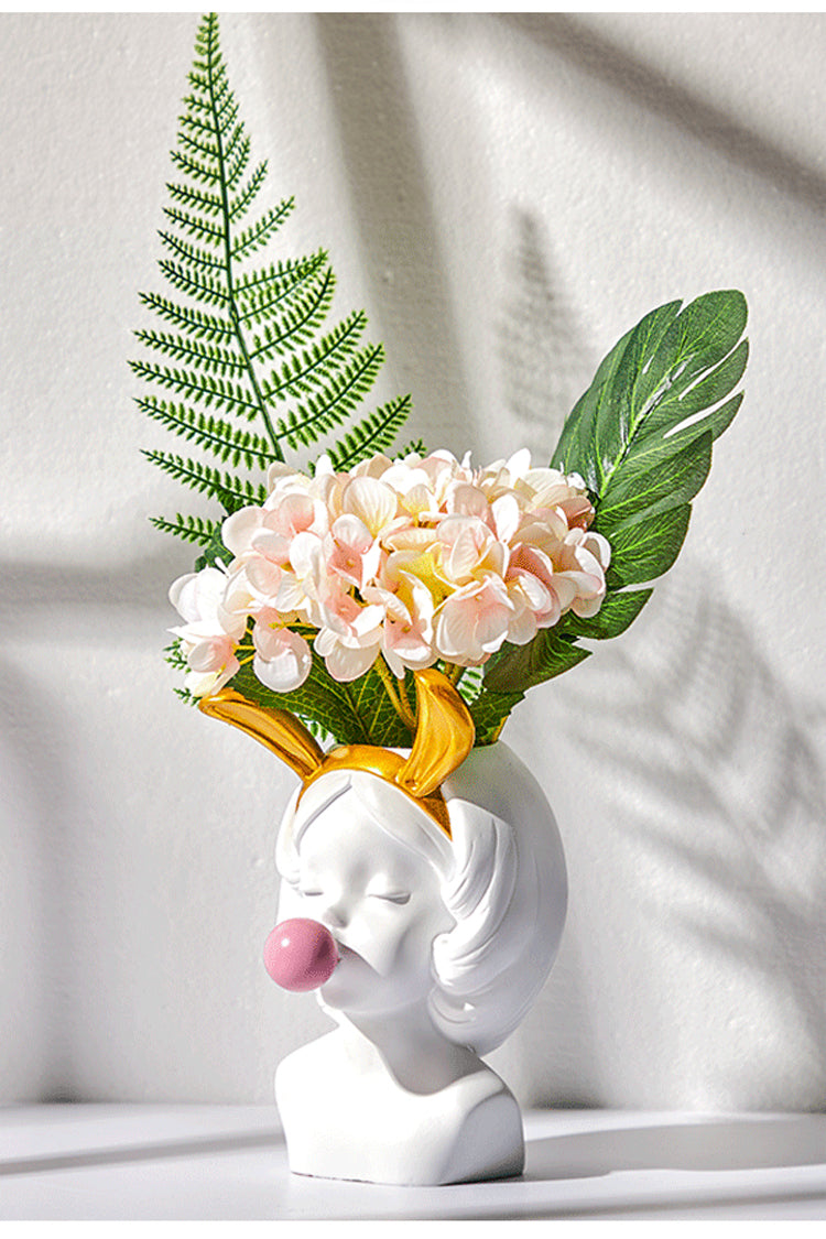 White girl head vase with gold bunny ears, blowing a pink bubble, holding pink flowers and green ferns.
