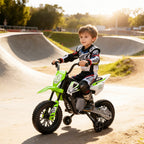 Child riding a green and white 12V electric motorcycle with training wheels on a dirt track at sunset.