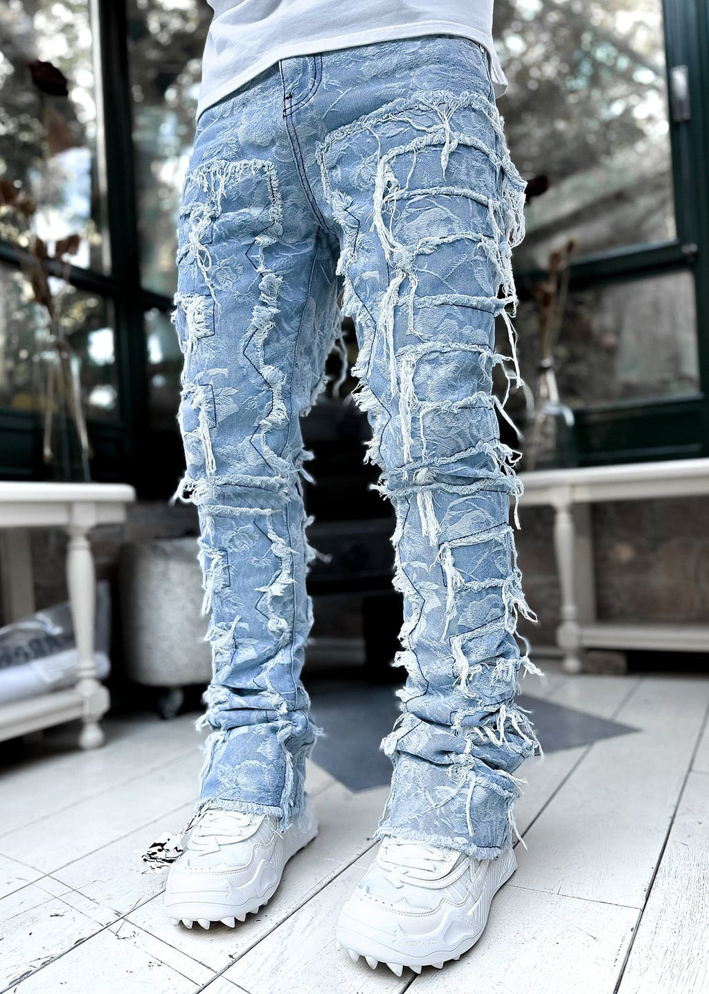 Light blue floral-patterned stacked jeans for men featuring distressed fringe patches and a slim, elongated fit.