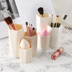 Beige, geometric plastic makeup brush holders of varying heights, containing brushes and beauty sponges.