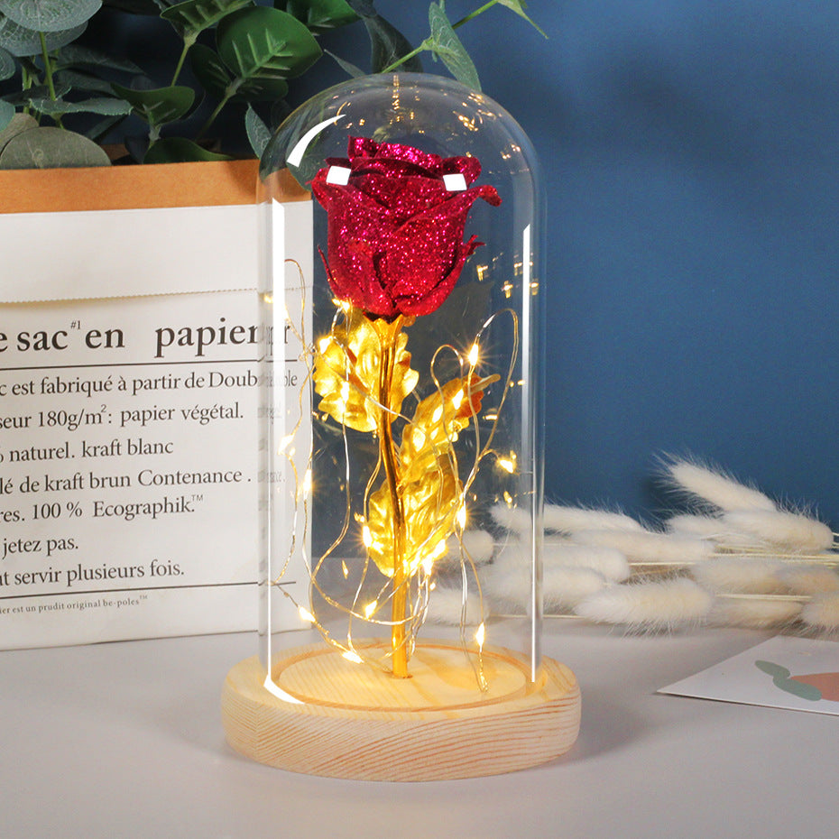 Glittery red rose with gold leaves inside a glass dome on a wooden base, illuminated by warm LED fairy lights.