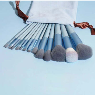 13-piece light blue makeup brush set with gray synthetic bristles, shown spilling from a matching fabric drawstring bag.