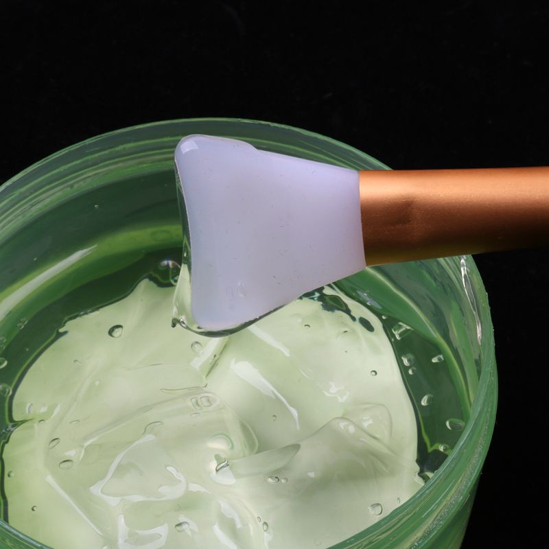 Silicone beauty mask brush with a bronze handle dipping into a container of translucent green skincare gel.